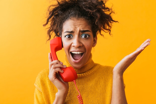 Foto retrato de uma mulher afro-americana chocada com um penteado afro, gritando e segurando o fone vermelho, isolado