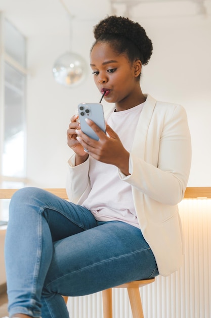 Foto retrato de uma mulher afro-americana mandando mensagens de texto para seus amigos no telefone inteligente sentado em uma cafeteria bebendo uma bebida tecnologia da internet retrato da beleza