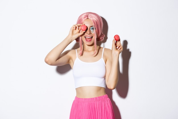 Retrato de uma mulher alegre e atraente com peruca rosa, comemorando o feriado, segurando macaroons e sorrindo, em pé sobre um fundo branco