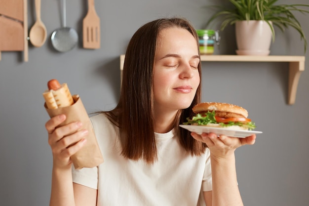 Retrato de uma mulher alegre e satisfeita com cabelos escuros, vestida com camiseta branca, segurando um grande sanduíche de hambúrguer e linguiça na massa, cheirando a aromat saboroso de junk food com fome