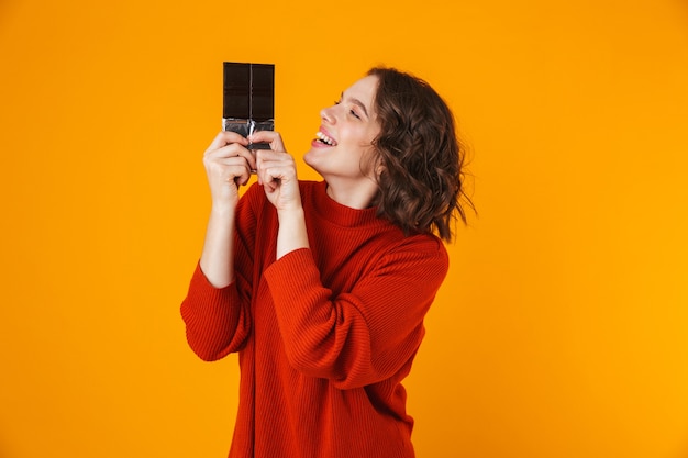 Retrato de uma mulher alegre vestindo um suéter, sorrindo e segurando a barra de chocolate em pé, isolado no amarelo