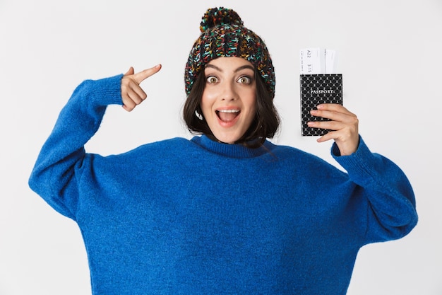 Retrato de uma mulher animada com chapéu de inverno segurando o passaporte e as passagens em pé, isolado no branco