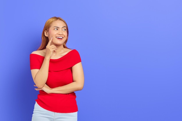 Retrato de uma mulher asiática sorridente feliz usando vestido vermelho olhando de lado com o dedo no queixo isolado em fundo roxo