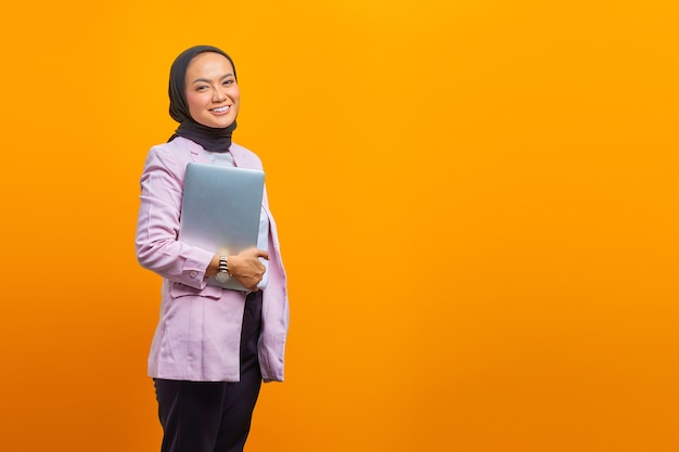 Retrato de uma mulher asiática sorridente segurando um laptop e olhando para a câmera sobre fundo amarelo