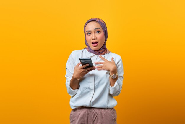 Retrato de uma mulher asiática surpresa usando um telefone celular com a boca aberta sobre um fundo amarelo