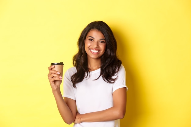 Retrato de uma mulher atraente afro-americana em camiseta branca, bebendo café e sorrindo em pé ov ...