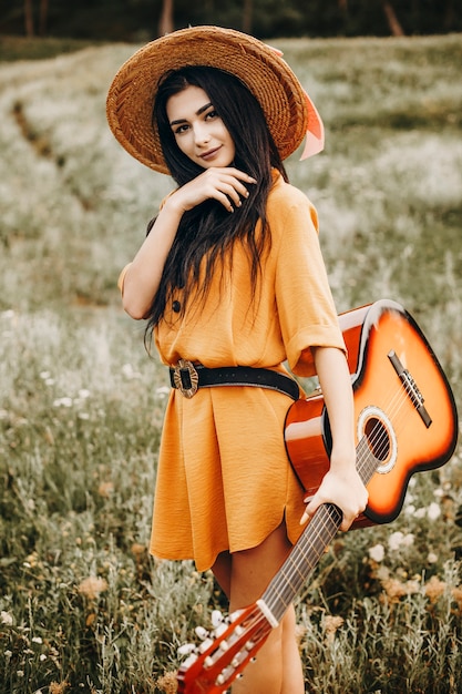 Foto retrato de uma mulher atraente caucasiana, segurando uma guitarra e olhando para a câmera, sorrindo contra uma colina com grama e flores.