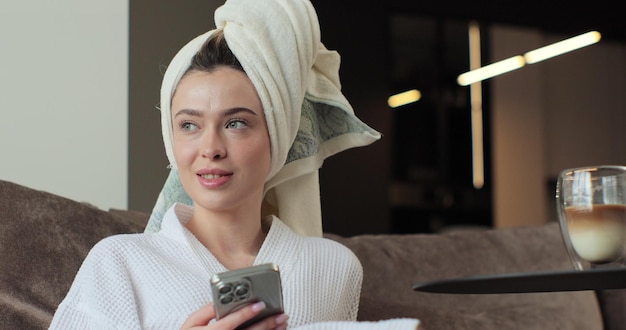 Foto retrato de uma mulher atraente depois de um banho vestindo um roupão de banho e uma toalha na cabeça está usando um smartphone no sofá na sala de estar relaxando depois de tomar um banho quente