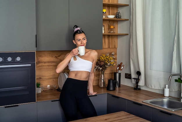 Retrato de uma mulher atraente segurando uma xícara e uma faca sorrindo em sua cozinha