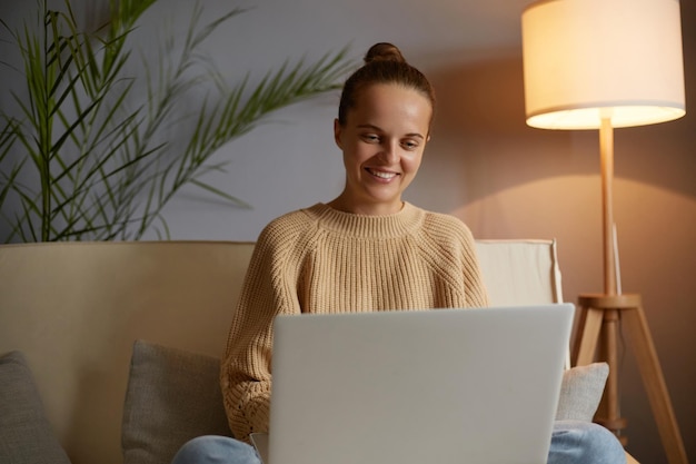 Retrato de uma mulher atraente sorridente com penteado de coque vestindo roupas casuais, sentado no sofá e trabalhando no laptop feminino satisfeito com seu trabalho olhando para a tela