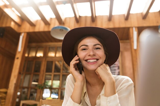 Retrato de uma mulher atraente usando chapéu, falando no smartphone, enquanto está sentado em um café aconchegante