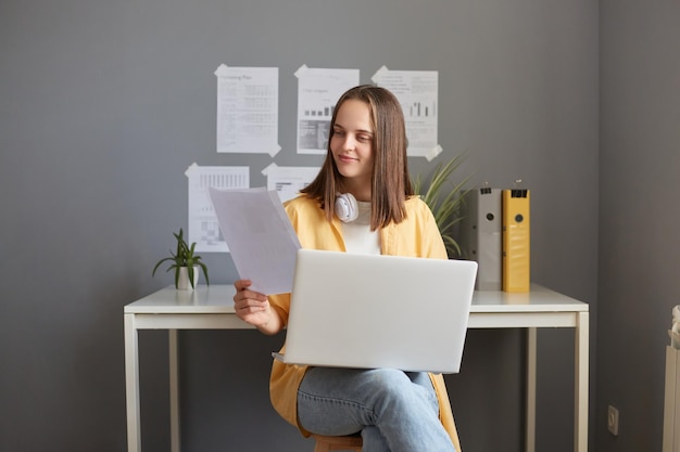 Retrato de uma mulher atraente vestindo jaqueta amarela sentada no local de trabalho e trabalhando em laptop lendo jornais fazendo seu trabalho expressando emoções positivas