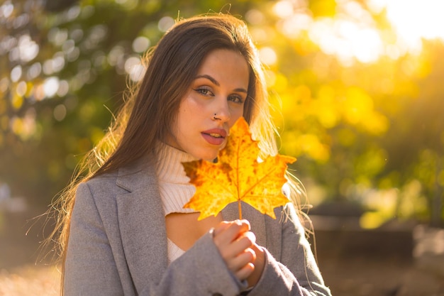 Retrato de uma mulher bonita aproveitando o outono em um parque ao pôr do sol com uma folha da árvore