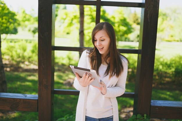 Retrato de uma mulher bonita com um sorriso encantador, vestindo roupas leves e casuais. Menina bonita segurando o computador tablet pc, lendo notícias no parque da cidade, na rua ao ar livre na natureza da primavera. Conceito de estilo de vida.