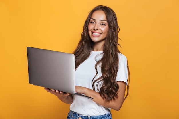 Retrato de uma mulher bonita de 20 anos com cabelo comprido, sorrindo e segurando um laptop cinza, isolado sobre uma parede amarela