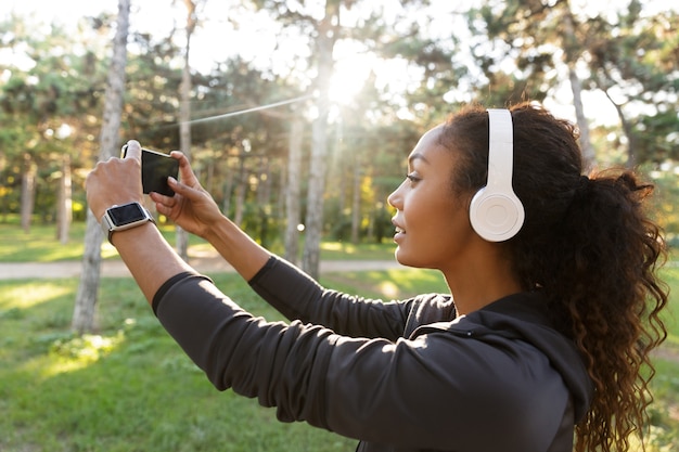 Foto retrato de uma mulher bonita de 20 anos usando um agasalho esportivo preto e fones de ouvido, tirando uma foto de selfie no celular enquanto caminha pelo parque verde