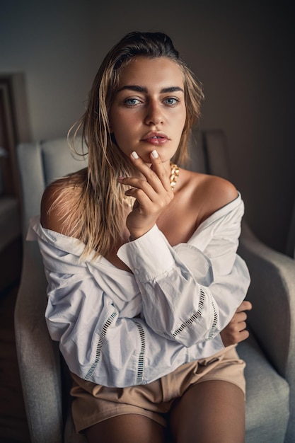 Retrato de uma mulher bonita e atraente com cabelo loiro sentado em uma cadeira em uma cadeira com uma camisa branca e posando lindamente