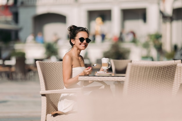 Retrato de uma mulher bonita em óculos de sol e vestido sentado em um café e bebendo café em um dia ensolarado de verão.