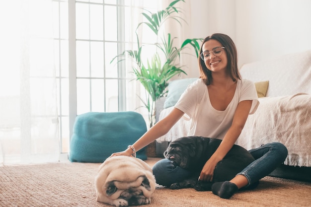 Retrato de uma mulher bonita em óculos se divertindo com seus cães de pugs de animais de estimação, sentados no chão da sala de estar de sua casa. Mulher alegre passando tempo de lazer com seus dois fofos cachorros de estimação em casa