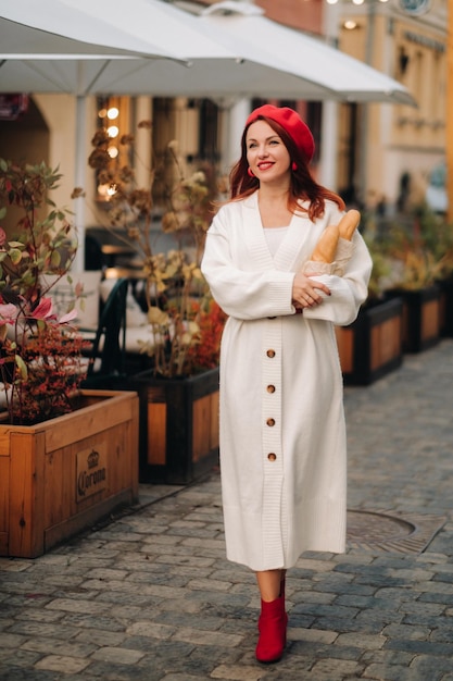 Retrato de uma mulher bonita em uma boina vermelha e um casaco de lã branco com baguetes nas mãos passeando pela cidade do outono