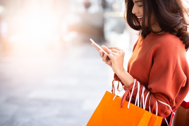 Retrato de uma mulher bonita feliz segurando sacolas de compras enquanto estiver usando o smartphone