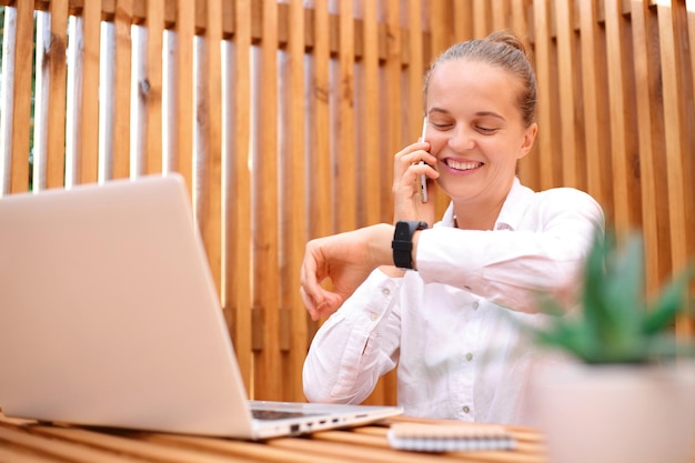 Retrato de uma mulher bonita sorridente vestindo camisa branca usando laptop enquanto está sentado no café e trabalhando no laptop falando no celular e olhando para sua reunião de panorâmica de relógio inteligente