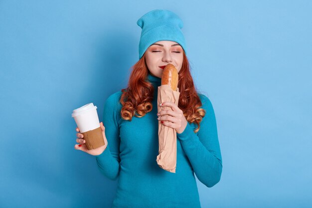 Retrato de uma mulher bonita vestindo um traje azul, sentindo o cheiro de pão fresco e segurando um café para viagem,