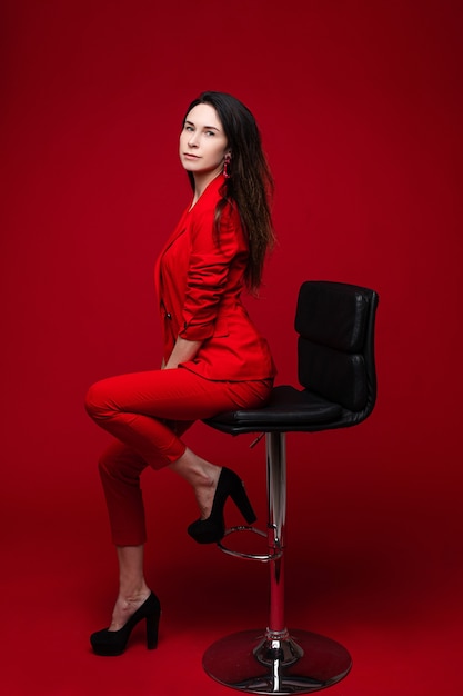 Retrato de uma mulher branca com cabelo longo e liso e escuro, em um terno vermelho, sapatos pretos, senta-se em uma cadeira preta