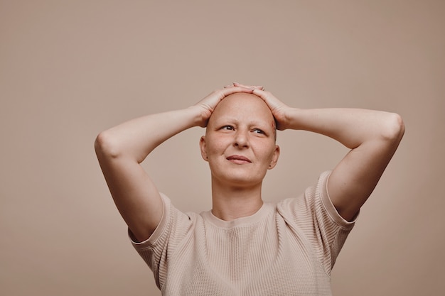 Foto retrato de uma mulher careca confiante em tons quentes olhando para longe com as mãos na cabeça enquanto posa contra um fundo bege mínimo em estúdio. alopecia e conscientização do câncer, copie o espaço