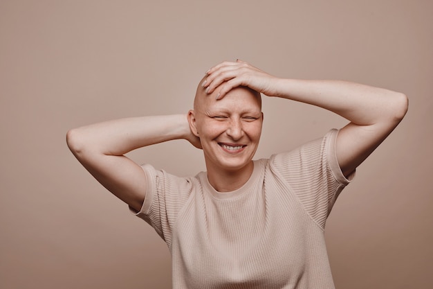 Retrato de uma mulher careca despreocupada em tons quentes tocando a cabeça raspada e sorrindo enquanto posava contra um fundo bege mínimo em estúdio. Alopecia e consciência do câncer, copie o espaço