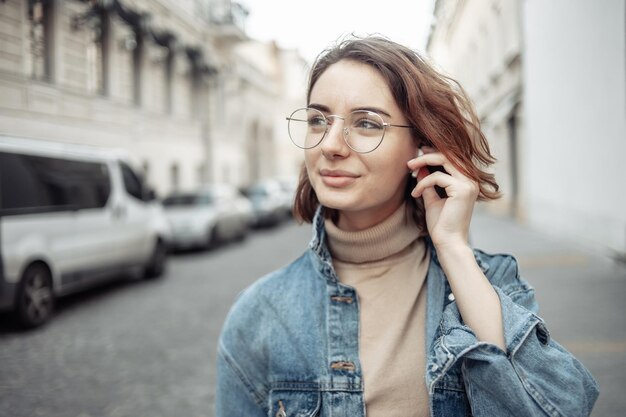 Retrato de uma mulher carismática elegante e moderna em roupas da moda com óculos e fones de ouvido Mulher jovem e atraente andando pela rua urbana