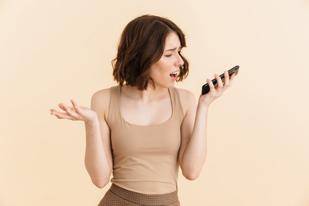 Retrato de uma mulher caucasiana descontente de 20 anos, vestida com roupas casuais, franzindo a testa enquanto faz uma chamada de celular no celular isolado