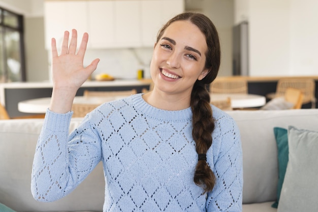 Retrato de uma mulher caucasiana feliz fazendo uma chamada de vídeo acenando com a mão e vestindo um suéter azul em casa. Vida doméstica, tecnologia e estilo de vida, inalterados.