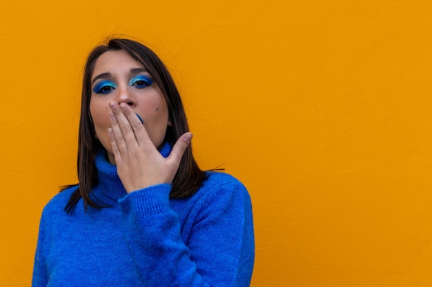 Retrato de uma mulher caucasiana vestida e composta de azul com um gesto surpreso com a mão na boca em um fundo amarelo