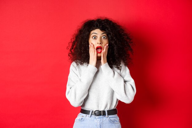 Retrato de uma mulher chocada grita de espanto, tocando o rosto e olhando para a câmera em uma impressionante oferta promocional, vestindo um moletom sobre fundo vermelho