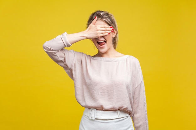 Retrato de uma mulher chocada ou assustada confusa com cabelos louros em pé de blusa cobrindo os olhos não quer assistir a expressão de medo ou vergonha tiro de estúdio interior isolado em fundo amarelo