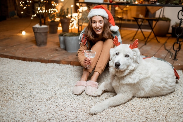 Retrato de uma mulher com chapéu de Natal com seu lindo cachorro comemorando o feriado de ano novo, sentados juntos no terraço lindamente decorado em casa