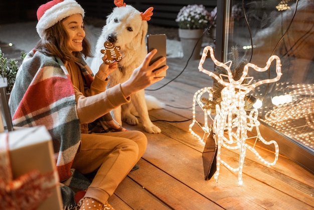 Retrato de uma mulher com chapéu de Natal e xadrez com seu cachorro fofo, comemorando as férias de ano novo em casa, alimentando o cachorro com biscoitos de gengibre e fazendo selfie uma foto