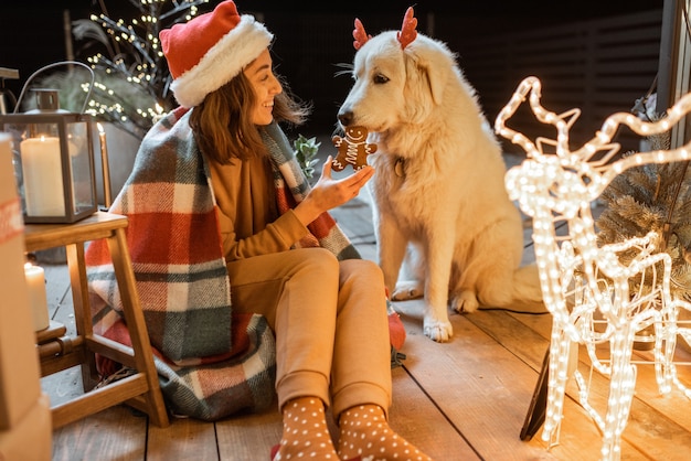 Retrato de uma mulher com chapéu de natal e xadrez com seu cachorro fofo, comemorando as férias de ano novo no terraço lindamente decorado de casa, alimentando o cachorro com biscoitos de gengibre