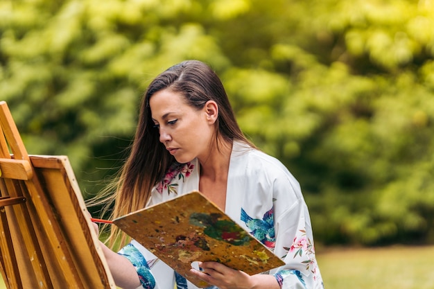 Retrato de uma mulher com expressão concentrada, pintando sobre tela em um parque.