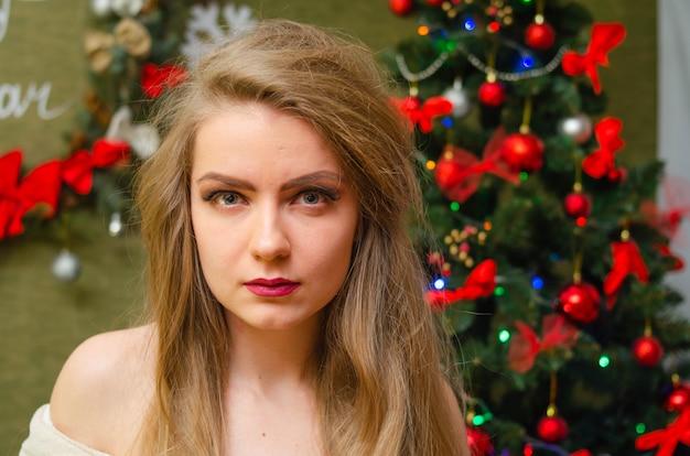 Retrato de uma mulher com lábios vermelhos brilhantes, cabelos longos loiros contra a árvore do ano novo. Jovem mulher com um casaco branco quente, os ombros são visíveis. Feriados. feliz Natal