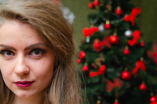 Retrato de uma mulher com lábios vermelhos brilhantes, cabelos longos loiros contra a árvore do ano novo. Jovem mulher com um casaco branco quente, os ombros são visíveis. Feriados. feliz Natal