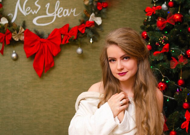 Retrato de uma mulher com lábios vermelhos brilhantes, cabelos longos loiros contra a árvore do ano novo. Jovem mulher com um casaco branco quente, os ombros são visíveis. Feriados. feliz Natal