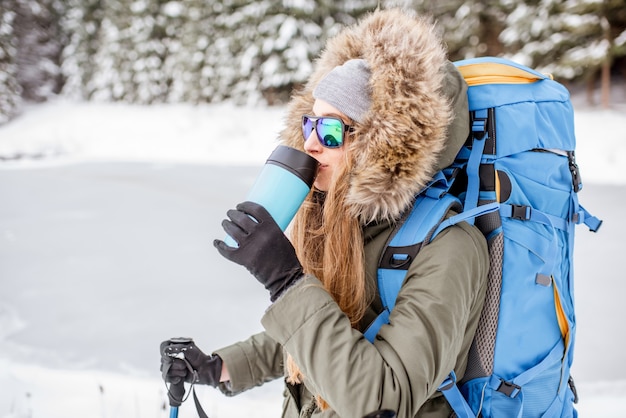 Retrato de uma mulher com roupas de inverno, caminhando com uma mochila, rastreadores e garrafa térmica no bosque nevado perto do lago congelado