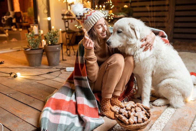 Retrato de uma mulher com seu cachorro fofo comemorando o feriado de ano novo, sentados juntos e comendo pão de gengibre no terraço lindamente decorado de casa