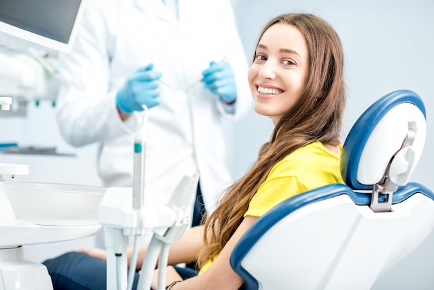 Retrato de uma mulher com sorriso dentuço sentada na cadeira odontológica com o médico no fundo do consultório odontológico