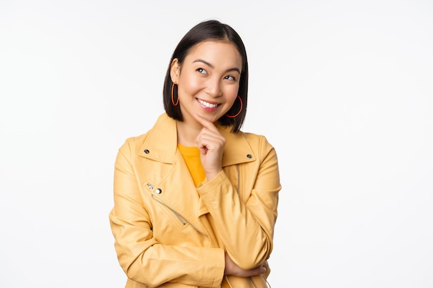Retrato de uma mulher coreana estilosa de jaqueta amarela sorrindo pensativo e olhando para o logotipo ou anúncio sobre fundo branco