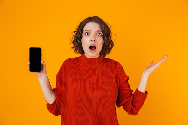 Retrato de uma mulher de 20 anos surpresa, vestindo um suéter, segurando e demonstrando o smartphone em pé, isolado sobre o amarelo
