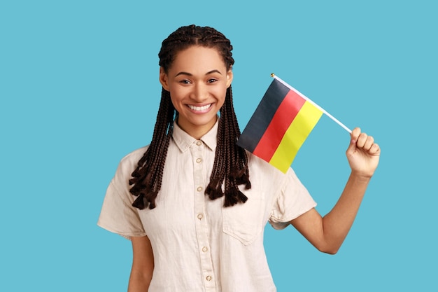 Retrato de uma mulher de aparência agradável com dreadlocks pretos segurando a bandeira da Alemanha comemorando o Dia da Alemanha 3 de outubro vestindo camisa branca Foto de estúdio interior isolada em fundo azul