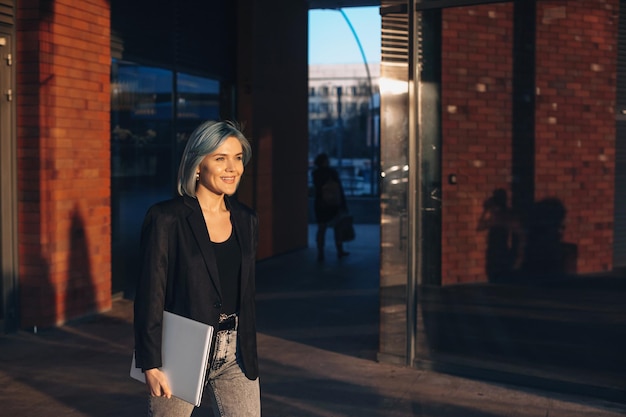 Retrato de uma mulher de cabelo azul segurando o laptop enquanto caminhava pela rua da cidade em um ensolarado